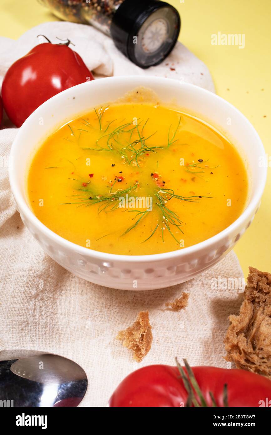 Weiße Schüssel mit Gemüsesuppe auf beigefarbener KüchenServiette mit Tomaten, Brot, Löffel und Gewürzen auf gelbem Hintergrund. Saisonale Ernährung. Sust Stockfoto