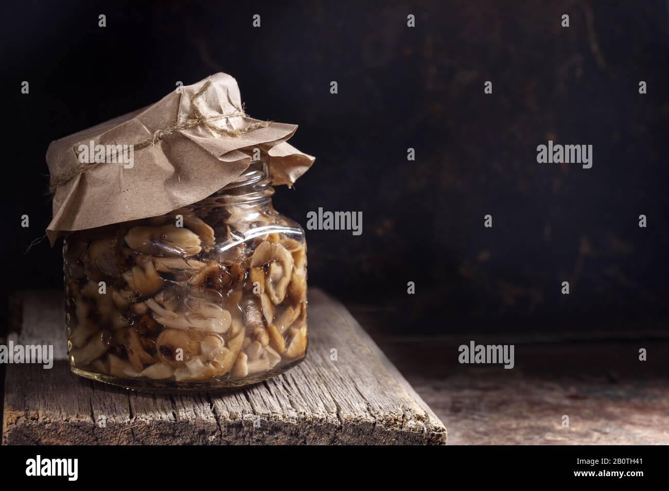 Glasbecher mit eingepickten Waldpilzen Honigagarika unter Papierabdeckung auf altem Holzbrett in dunkler rostiger Metallkulisse. Fermentierte gesunde Lebensmittel. Sust Stockfoto