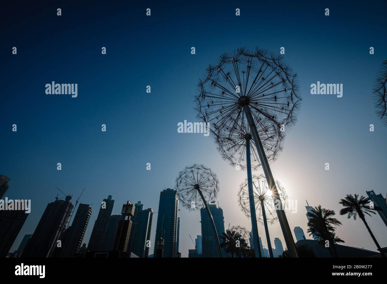 Riesige Metall-Löwenzahn-Skulptur im A-Park in Dubai, Silhouette von Löwenzahn und Wolkenkratzern gegen einen strahlenden Himmel bei Sonnenuntergang Stockfoto