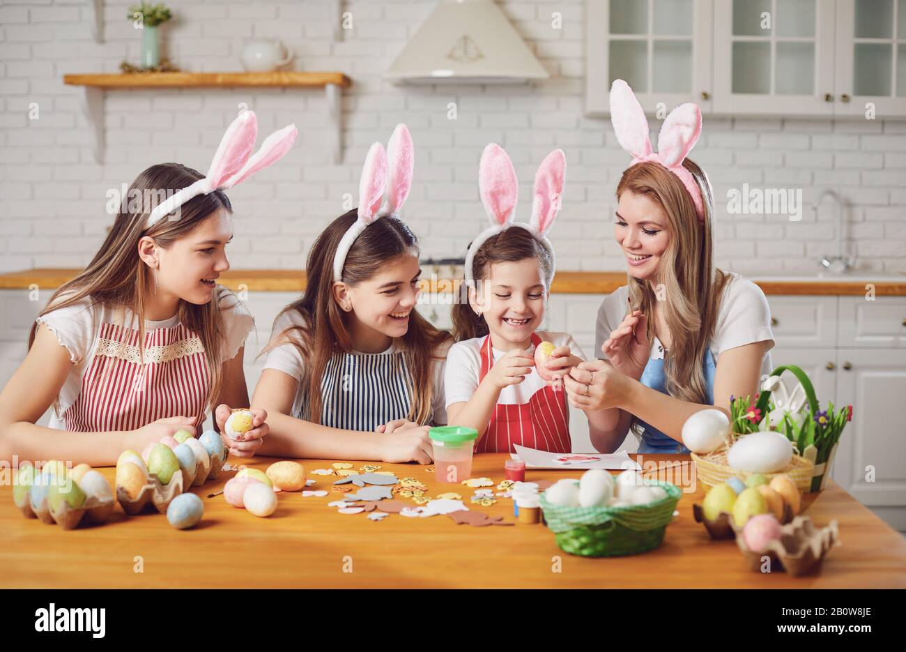 Frohe ostern. Mutter und Töchter mit Kaninchenohren schmücken Ostereier an einem Tisch in der Küche. Stockfoto