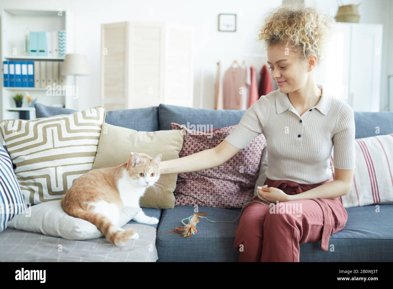 Junge Frau in legerer Kleidung, die während ihres Spiels zu Hause auf dem Sofa sitzt und Hauskatze streichelt Stockfoto