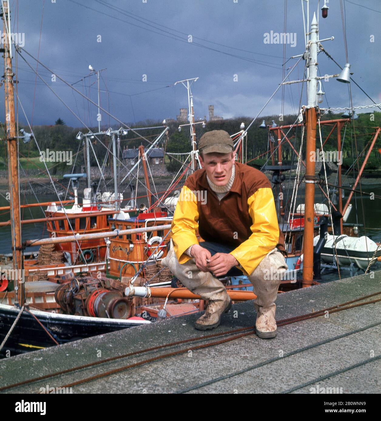 1960er Jahre, historisch, ein junger schottischer Fischer in seiner wasserdichten Angelkleidung und Kappe, kniend für ein Foto am Hafen von Stornoway, Isle of Lewis, Scottish Highland, Schottland, Großbritannien. Stockfoto