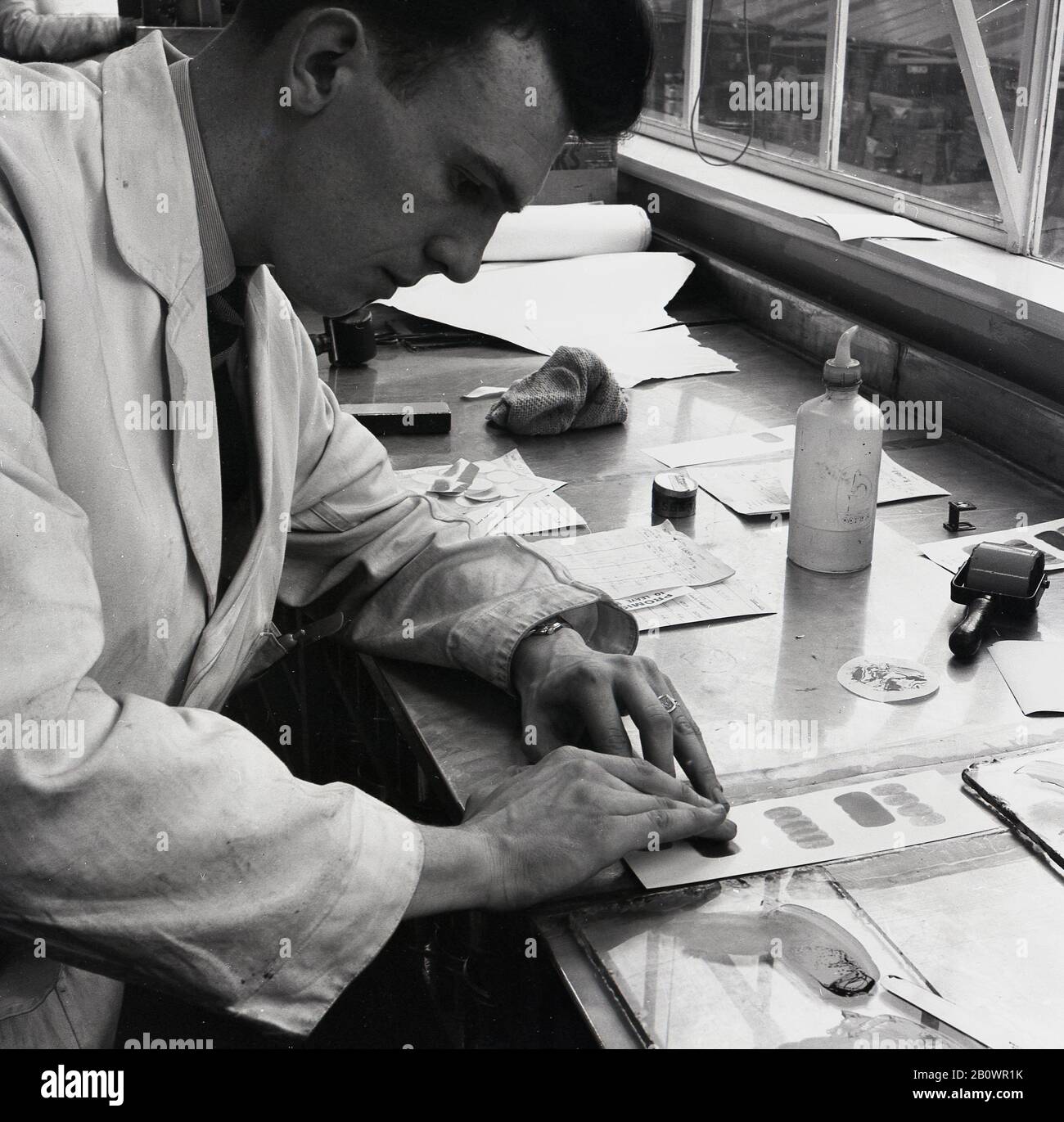 1950er Jahre, historisch, ein männlicher Techniker in einem weißen Mantel, der an einem Fenstertisch in einer Lackiererei arbeitete und mit den Fingern Farbe auf Papier mischte, um Farbe zu überprüfen, England, Großbritannien. Stockfoto