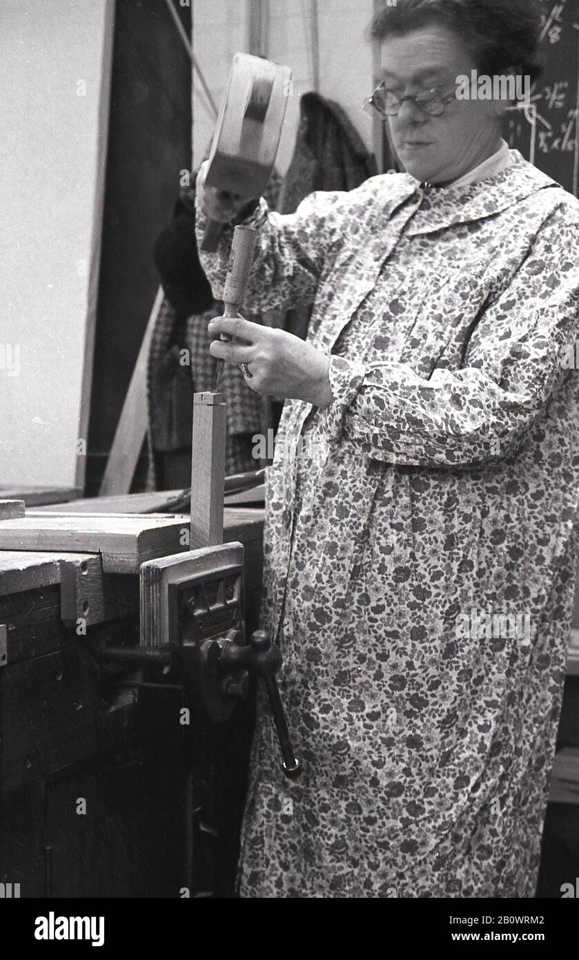 40er Jahre, historisch, Dame in einer Holzarbeitsklasse, mit  Blumenpattner-Schürze auf einer Werkbank mit Holzhammer und Meißel, um ein  Holzstück zu machen, das in einem Metall-Schraubstock befestigt ist,  England UK Stockfotografie - Alamy