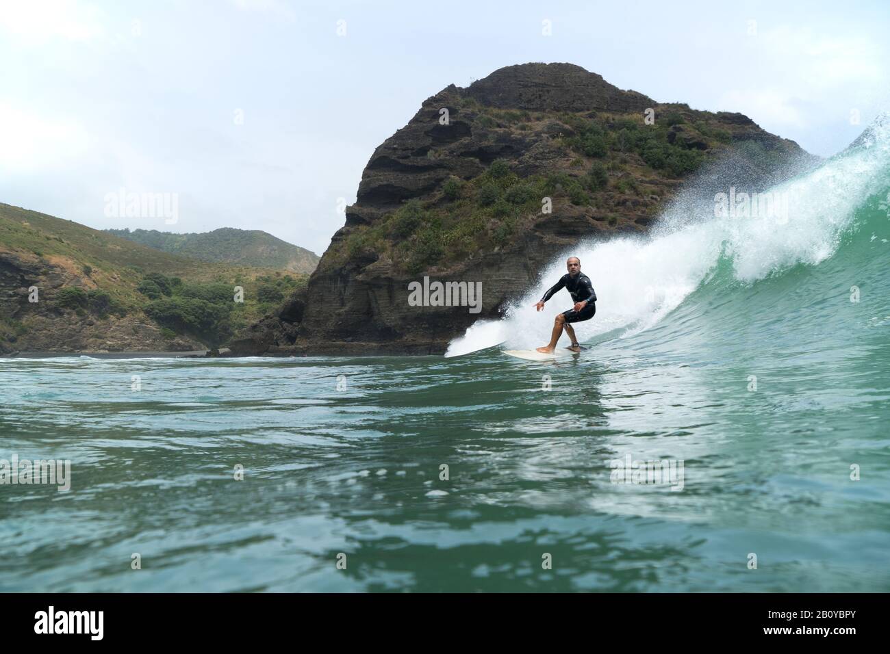 Ein Mann mittleren Alters surft auf einer großen grünen Bruchwelle am Piha Beach, Auckland, Neuseeland. Felsige Küste im Hintergrund, blauer Himmel mit Wolken. Stockfoto