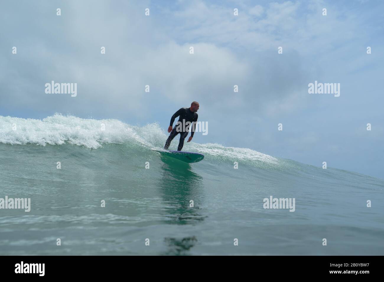 Ein junger männlicher Surfer reitet eine grüne Bruchwelle bei Piha, Neuseeland. Blauer Himmel mit Wolken. Stockfoto