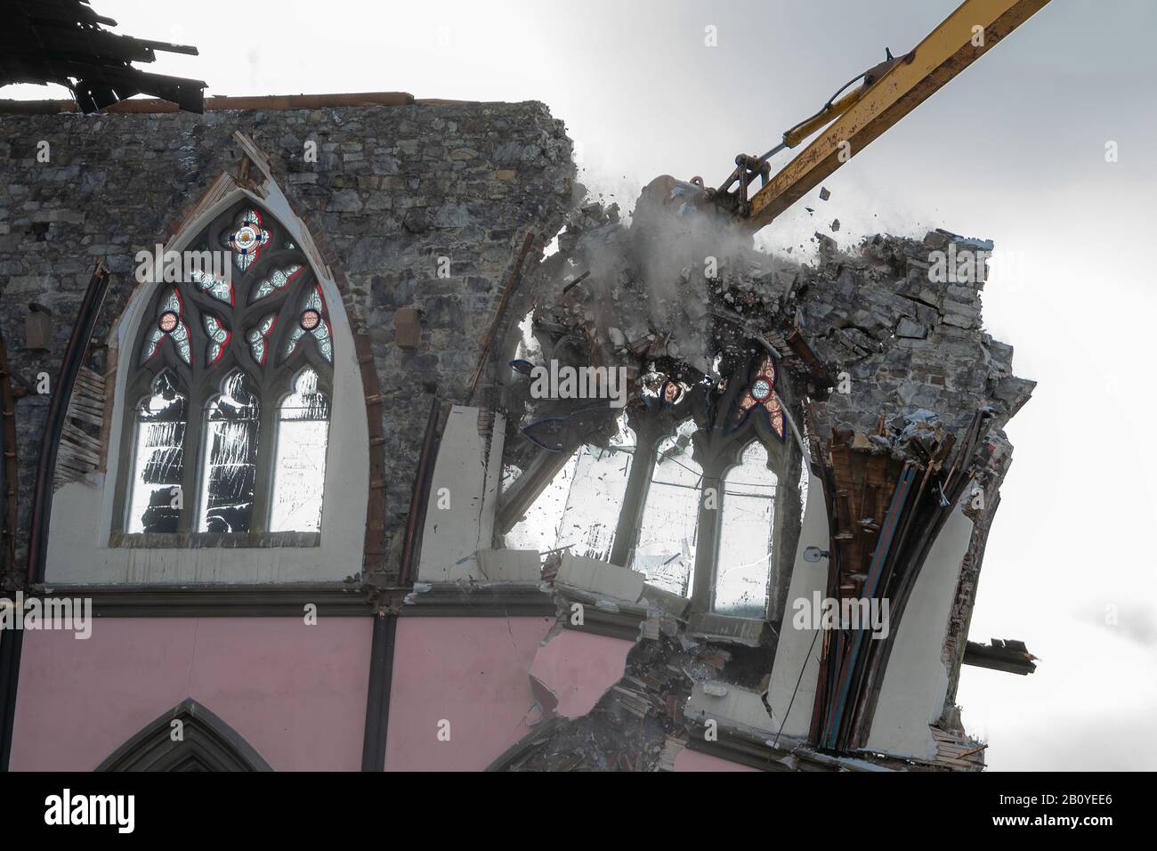 Action-Schuss eines großen Baggerladers, der eine gothische Kirche abreißen lässt. Trümmer fliegen um eine Wand und ein Fenster, während die Maschine sie herunterzieht. Übergiebeltem Himmel. Stockfoto