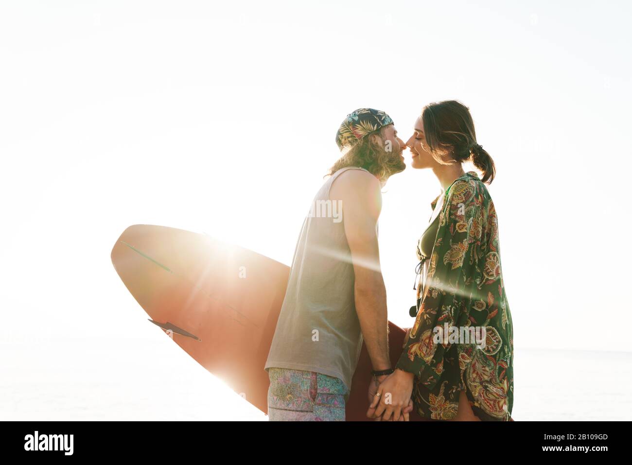Foto eines netten romantischen Ehepaares, das Surfbrett hält und beim laufen am sonnigen Strand küsst Stockfoto