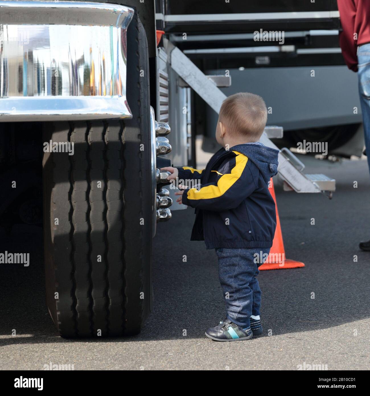 Kleiner Kleinkind in einer blauen Jacke, Hose und Stiefel neben einem großen Vorderrad eines Staplers, das Chrom-Tassenknüsse am Rad berührt. Stockfoto