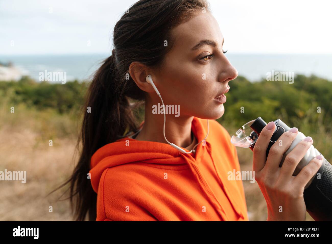 Foto einer netten Sportlerin in Sportbekleidung mit Ohrhörer und Trinkwasser während der Arbeit an der Küste Stockfoto