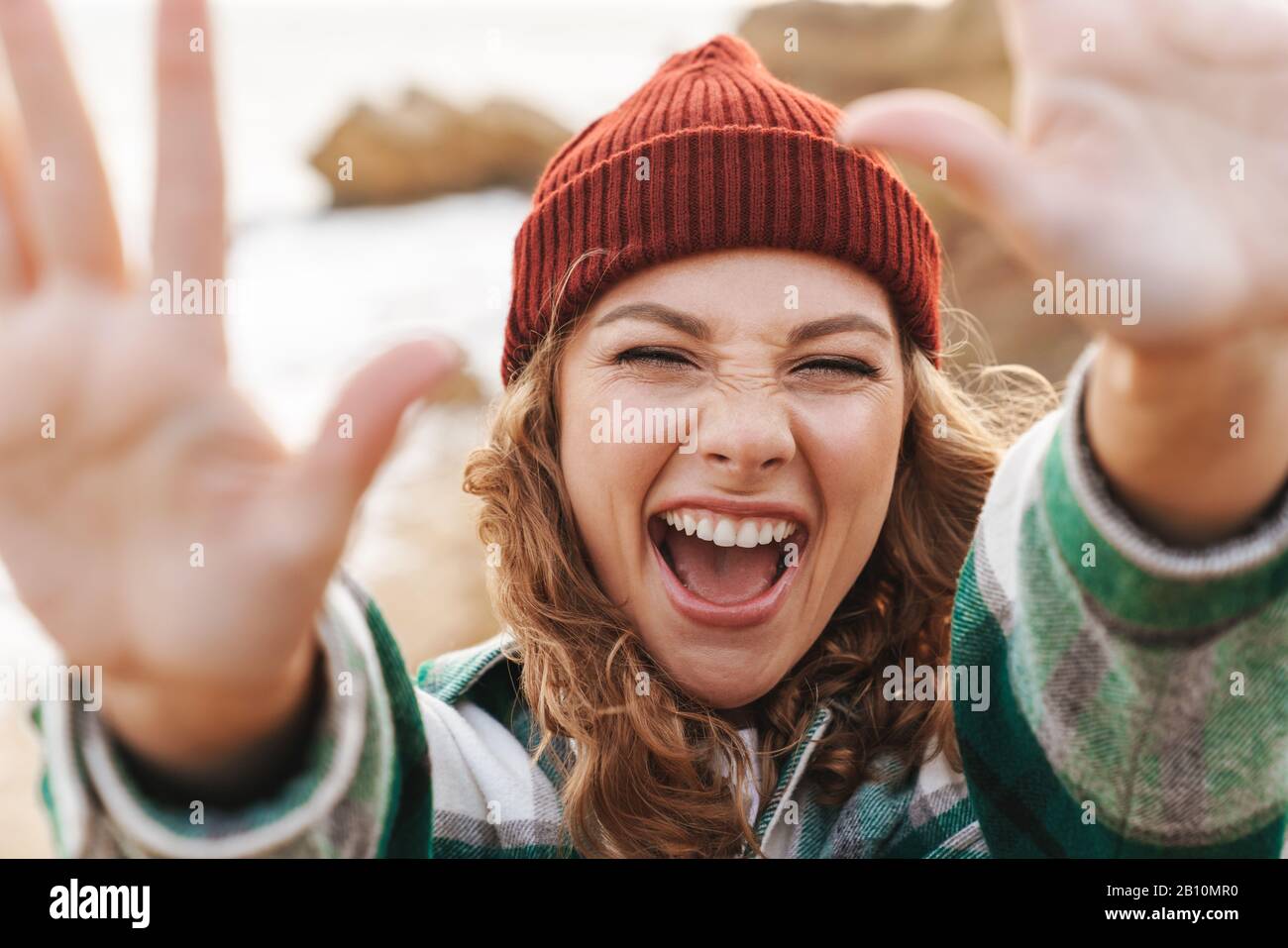Bild der freudigen jungen kaukasischen Frau, die Hut und ein Plaid Hemd trägt, das die Hände bei der Kamera erreicht, während sie im Freien spazieren geht Stockfoto