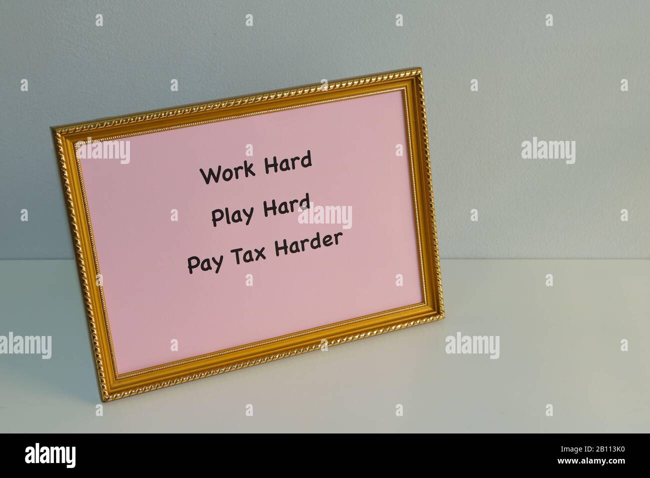 Close-Up-Slogan Hart Arbeiten, hart spielen, zahlen Steuern Härter gedruckt auf pinkfarbenem Papier in goldenem Fotorahmen auf weißem Holztisch Stockfoto