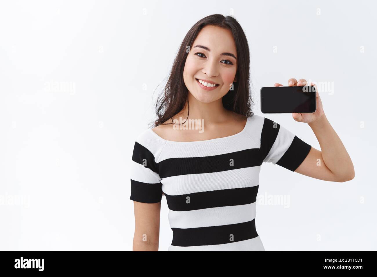 Fröhliche junge asianerin im gestreiften T-Shirt, das das Smartphone horizontal in der Nähe des Gesichts hält, den Kopf neigen und lächelnd freundlich lächeln, empfehlen Sie die App Stockfoto