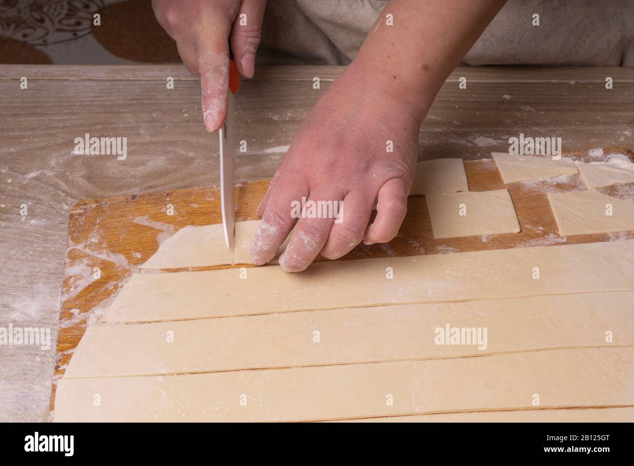 Eine Frau zerschneidet den Teig mit einem weißen Keramikmesser in Quadrate. Sperrholzschneidplatte, Holzmehlsieb und Walzstift aus Holz - Werkzeuge für die Herstellung Stockfoto