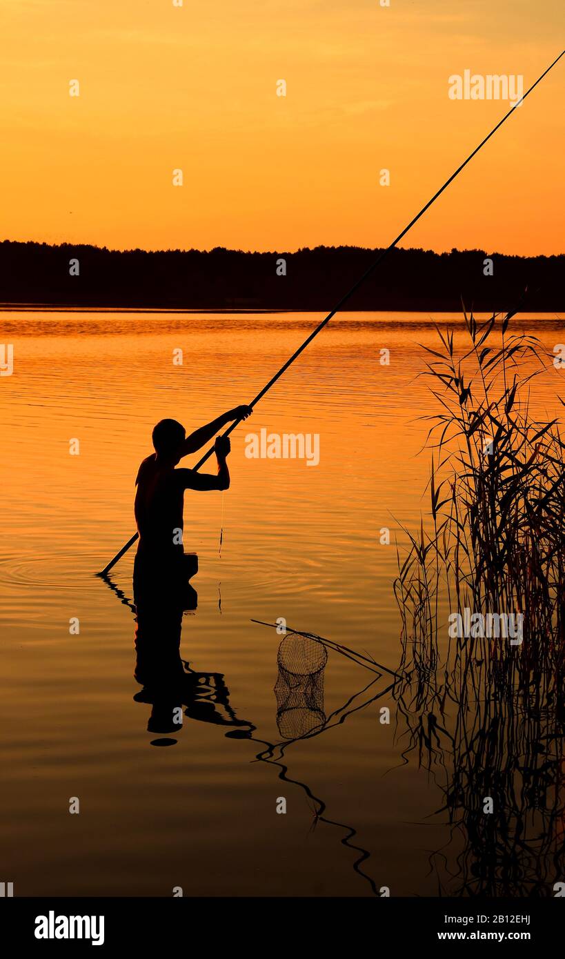 Dunkle Silhouette eines Mannes, der mit Angelrute über lebhaftem orangefarbenem Himmel und Hintergrund des Sees im Wasser steht, Rückansicht Stockfoto