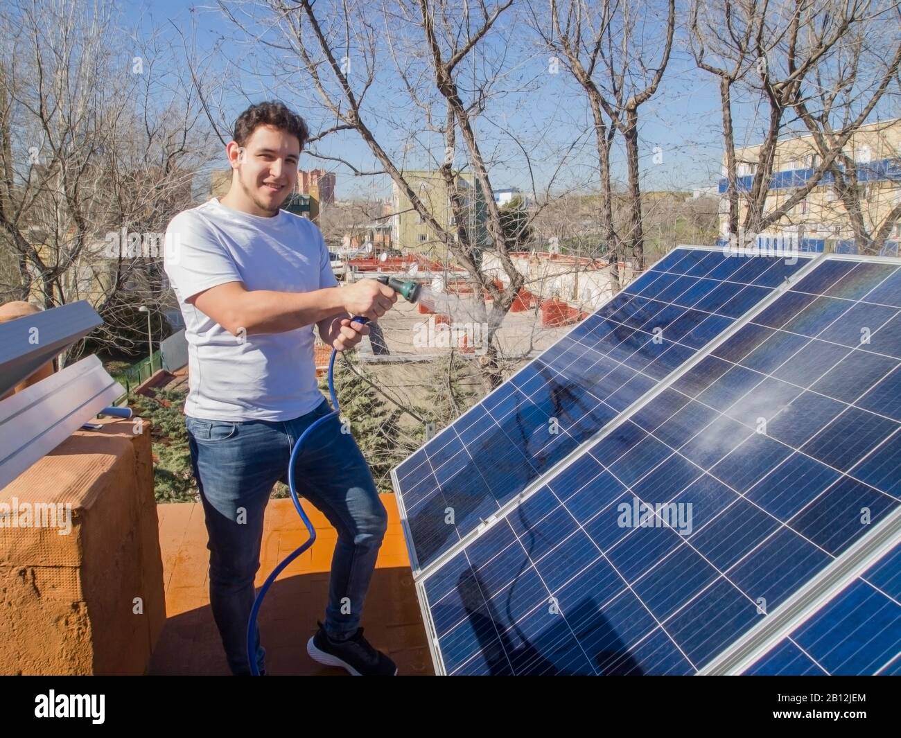 Kaukasischer junger Mann lächelt und säubert Solarpaneele mit einem Schlauch. Alternative Stromquelle, nachhaltige Ressourcenkonzeption. Stockfoto