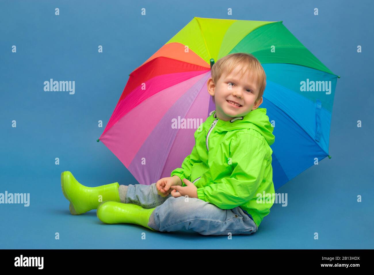 Ein kaukasisches Kind sitzt in einer Windschutzjacke und Gummistiefel unter einem Regenschirm - einem Regenbogen, auf blauem Hintergrund. Konzept zu Wetter und Jahreszeiten Stockfoto