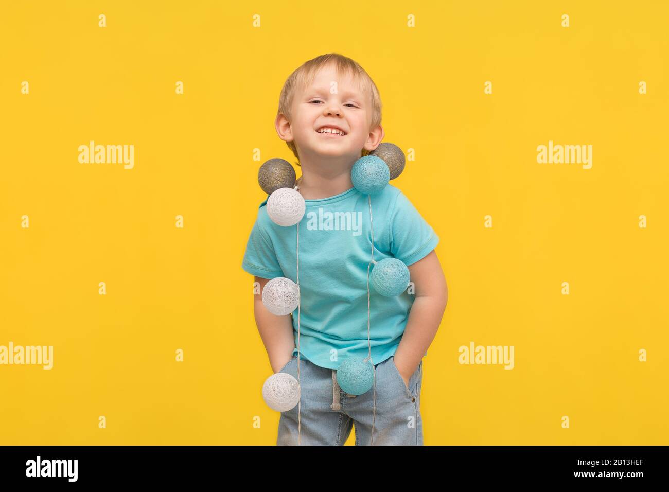 Fröhliches blondes Knabenkind in einem blauen T-Shirt lacht auf gelbem Grund, und als Dekoration hängt ihm eine Girlande am Hals. Das Konzept der Kindheit und p Stockfoto