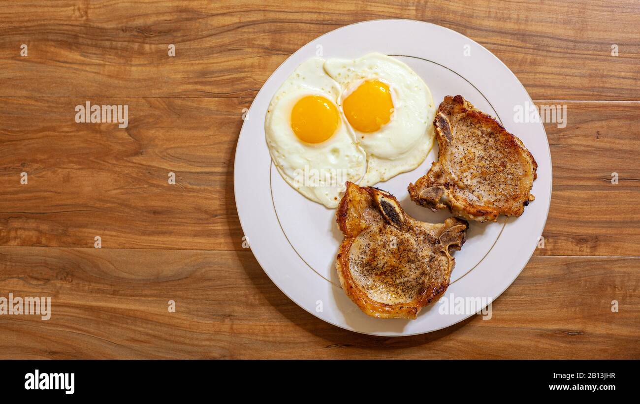 Draufsicht auf das Frühstücksgericht mit sonnigen Spiegeleiern und Pfannenfrittierten Schweinekoteletts, gewürzt mit schwarzem Pfeffer auf Holztisch. Stockfoto