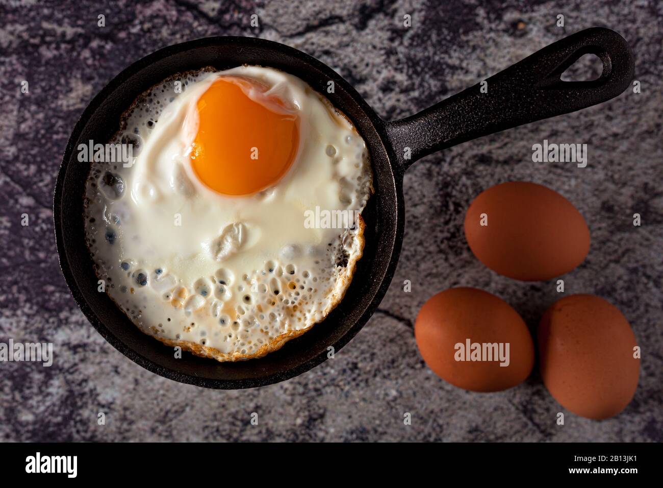 Sonnige Seite nach oben gebratenes Ei in Gusseisen-Skillet mit ganzen Eiern auf Betongrund. Draufsicht Stockfoto