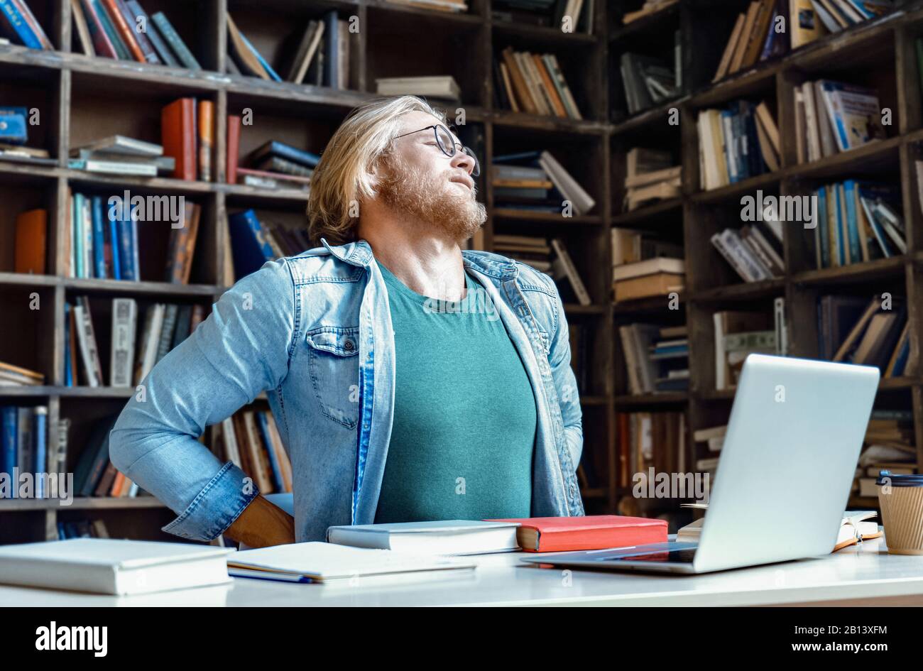 Müde aufgebrachten jungen Mann Brille Stretch Bibliothek Schreibtisch spüren Rückenschmerzen Konzept Stockfoto