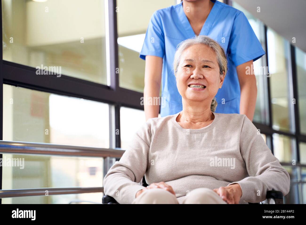 Asiatische Pflegekraft unterstützt Rollstuhlgebundene Seniorin im Flur des Pflegeheims Stockfoto