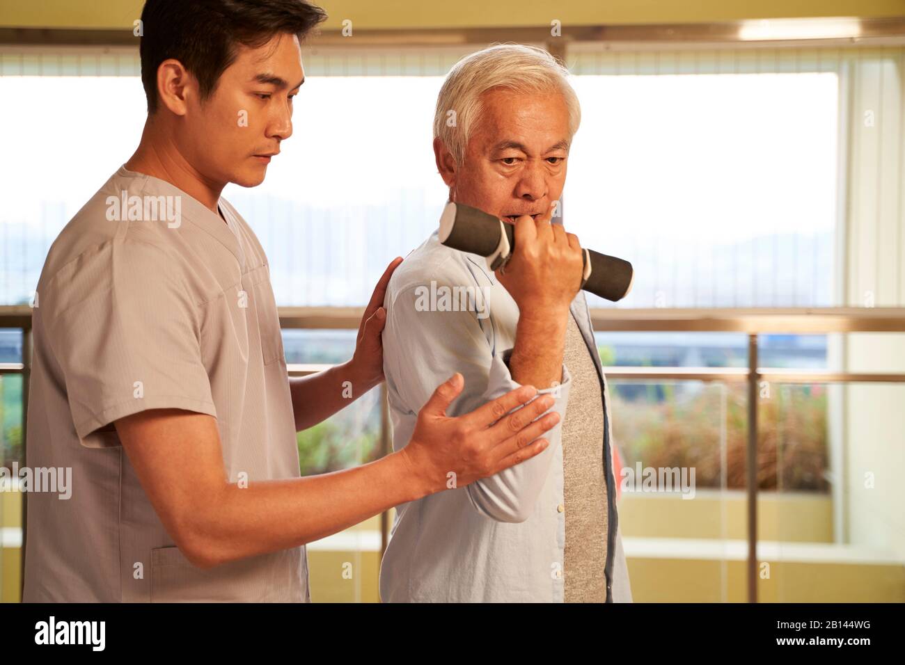Leitender asiatischer Mann, der mit Hanteln trainiert, die von einem physischen Therapeuten im Rehabilitationszentrum geleitet werden Stockfoto