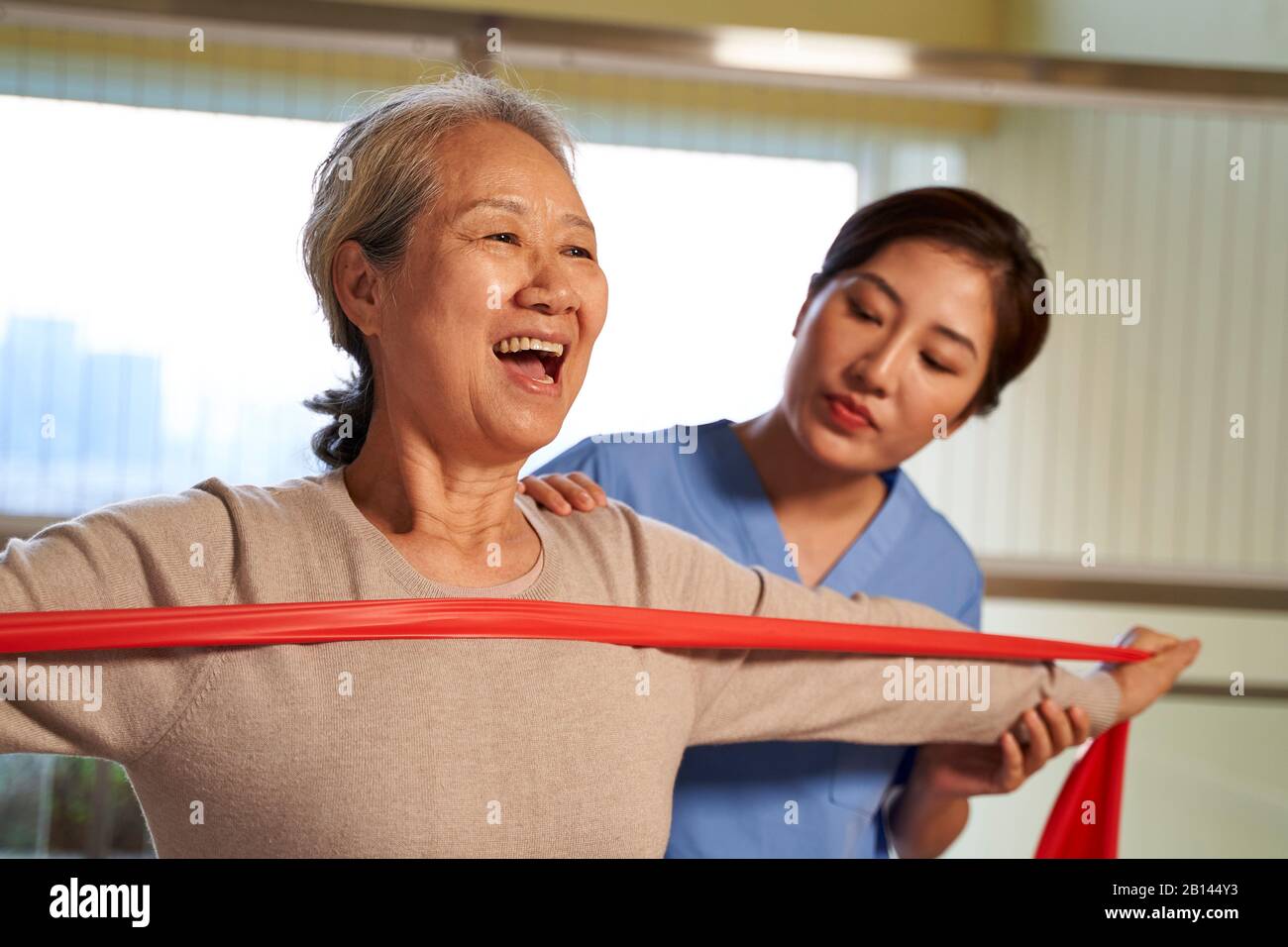 Glückliche ältere asiatische Frau, die mit einer Widerstandsband trainiert, die von einem physischen Therapeuten im Reha-Zentrum geleitet wird Stockfoto