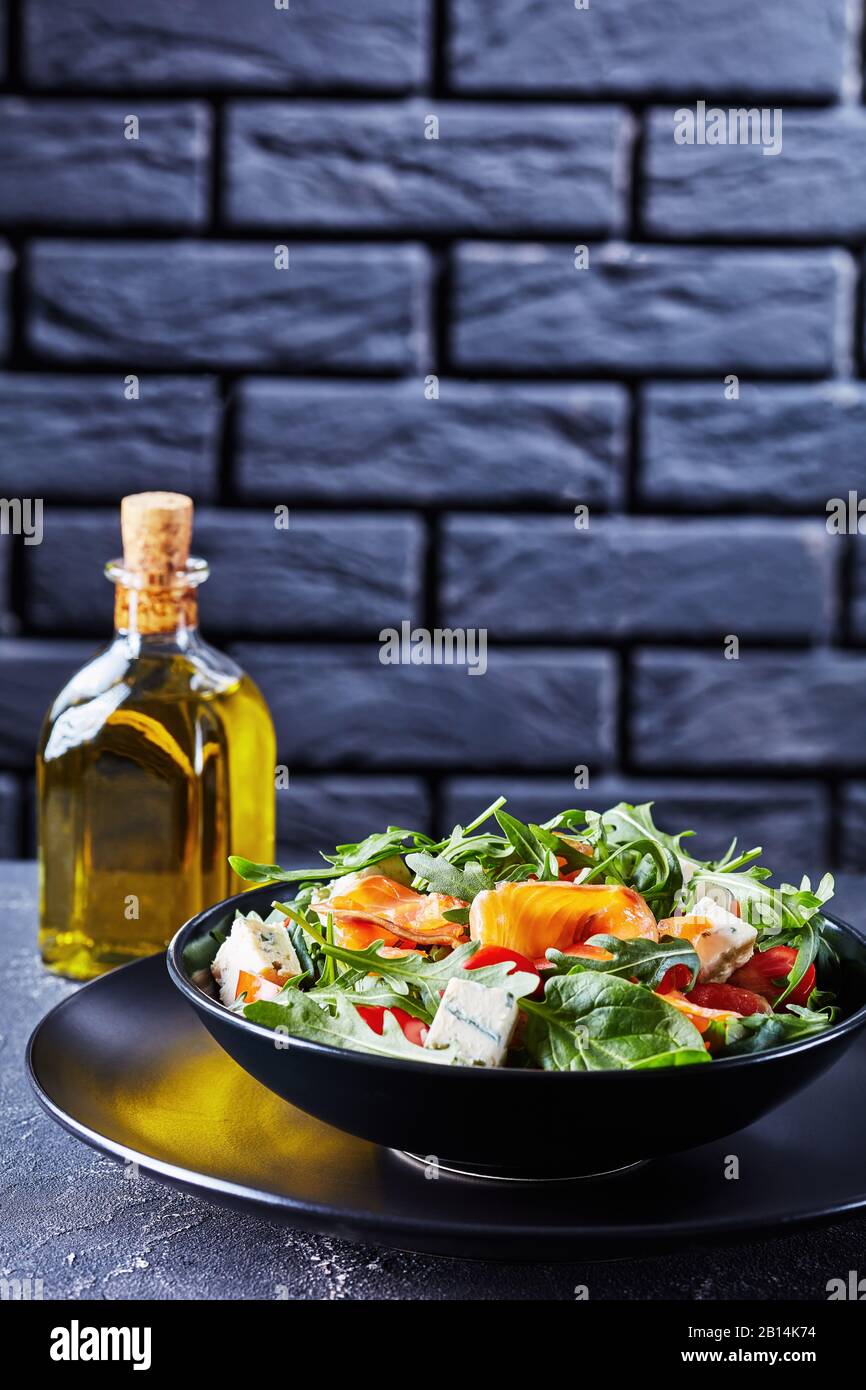 Nahaufnahme von köstlichem gesundem Salat aus Lachsarugula-Blaukäse-Tomaten in einer schwarzen Schüssel mit dunkler Ziegelwand im Hintergrund, vertikaler Blick von Abo Stockfoto