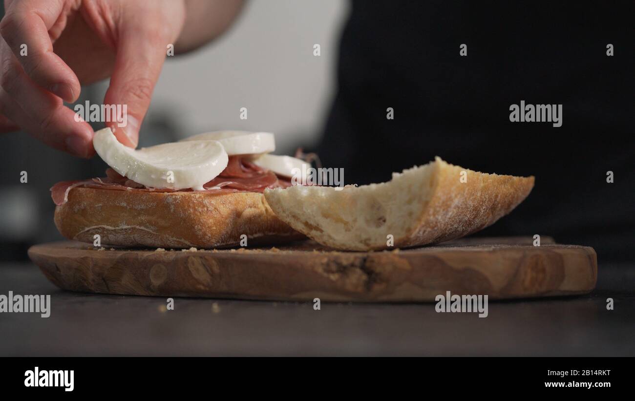 Man fügt Mozzarella auf Ciabatta auf olivem Brett hinzu, breites Foto Stockfoto