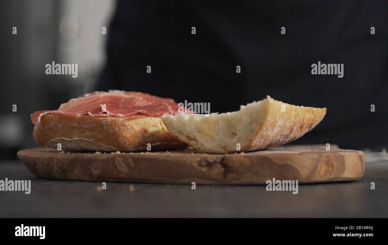 Man fügt Prosciutto auf Ciabatta auf olivem Brett hinzu, breites Foto Stockfoto