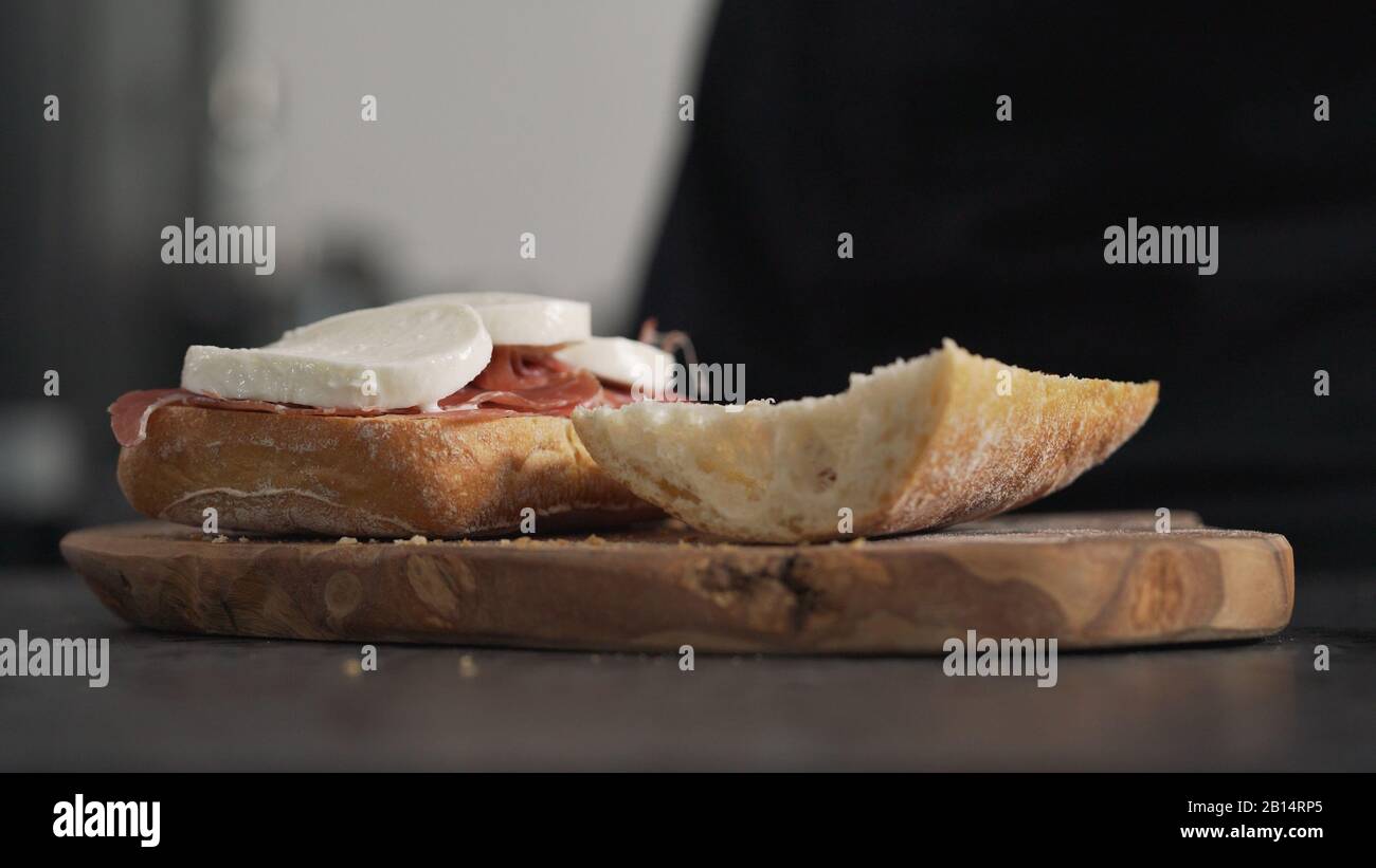 Man fügt Mozzarella auf Ciabatta auf olivem Brett hinzu, breites Foto Stockfoto