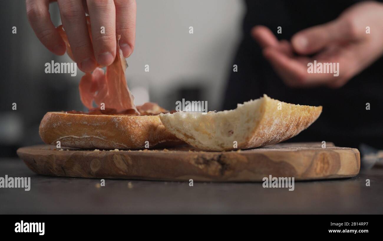 Man fügt Prosciutto auf Ciabatta auf olivem Brett hinzu, breites Foto Stockfoto
