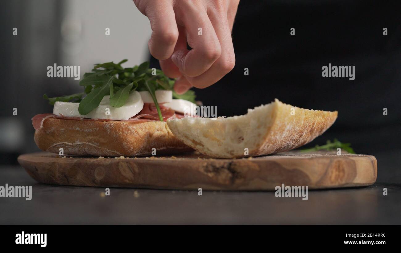 Man fügt auf Ciabatta auf olivem Brett eine Arugula hinzu, ein breites Foto Stockfoto