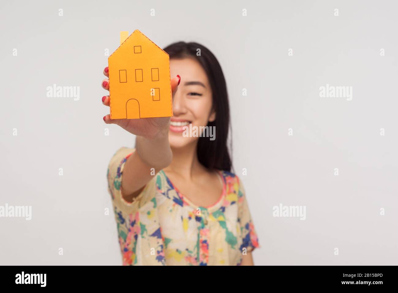 Immobilienkauf, zuverlässige Immobilienagentur. Fröhliches asiatisches Mädchen mit brünettem Haar in der Sommer-Bluse, das Papierhaus in ausgestreckter Hand zeigt Stockfoto