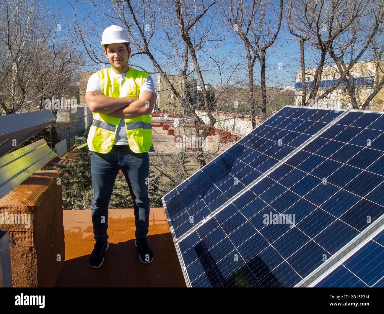 Kaukasisch attraktiver junger Techniker mit durchgestrichenen Armen und Blick auf die Kamera neben den Sonnenkollektoren. Alternative Stromquelle, nachhaltig Stockfoto