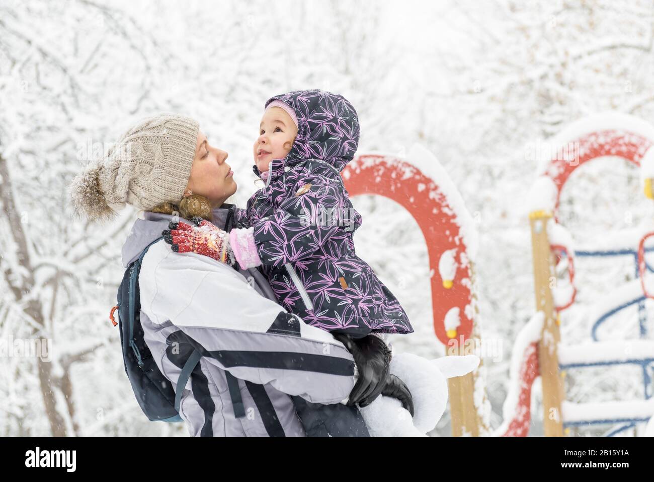 Fröhliches kleines Kind spielt im Winter mit der Mutter in einem verschneiten Park. Fröhliche Familienspaziergänge bei Schneefall. Süßes Baby-Mädchen sitzt auf den Händen der Mutter. Stockfoto