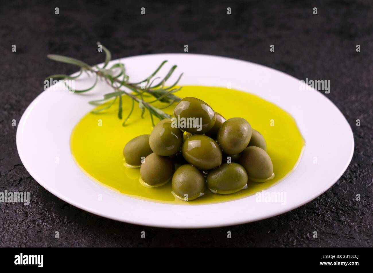 Oliven werden mit Olivenöl und einem Rosmarinspring auf einem weißen Teller gegossen. Das Konzept des Olivenöls. Stockfoto