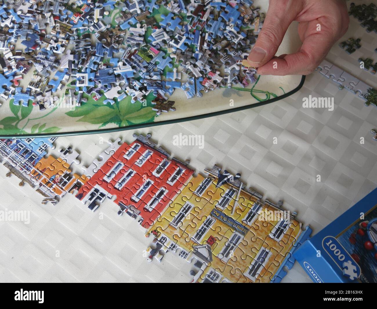 Ein Tablett mit Puzzleteilen und einer Hand, die ausgestreckt ist, um ein weiteres Stück in das Puzzle zu passen, das auf dem Esstisch gelegt ist. Stockfoto