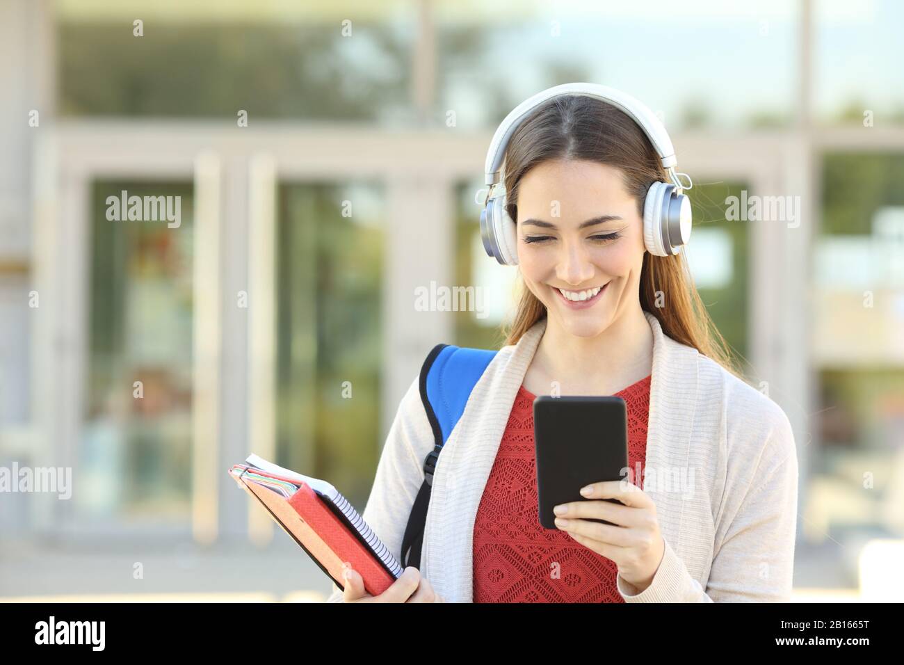 Vorderansicht Porträt eines glücklichen Studenten, der drahtlose Kopfhörer trägt, Musik mit dem Handy auf einem Campus anhört Stockfoto