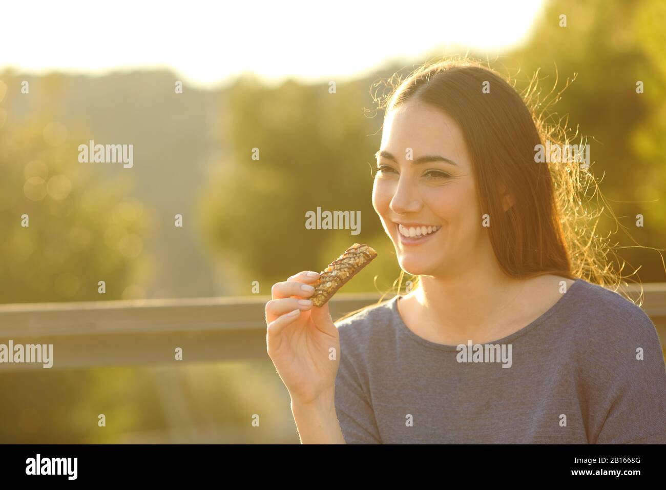 Fröhliche Frau, die bei Sonnenuntergang in einem Park eine Getreidebar auf einer Bank isst Stockfoto