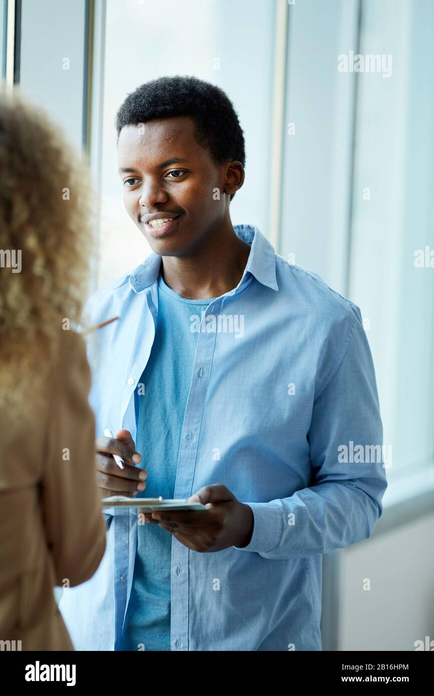 Vertikales Porträt eines jungen afroamerikanischen Mannes, der mit einer Freundin spricht, während er am Fenster in der Hochschule oder im Büro steht Stockfoto