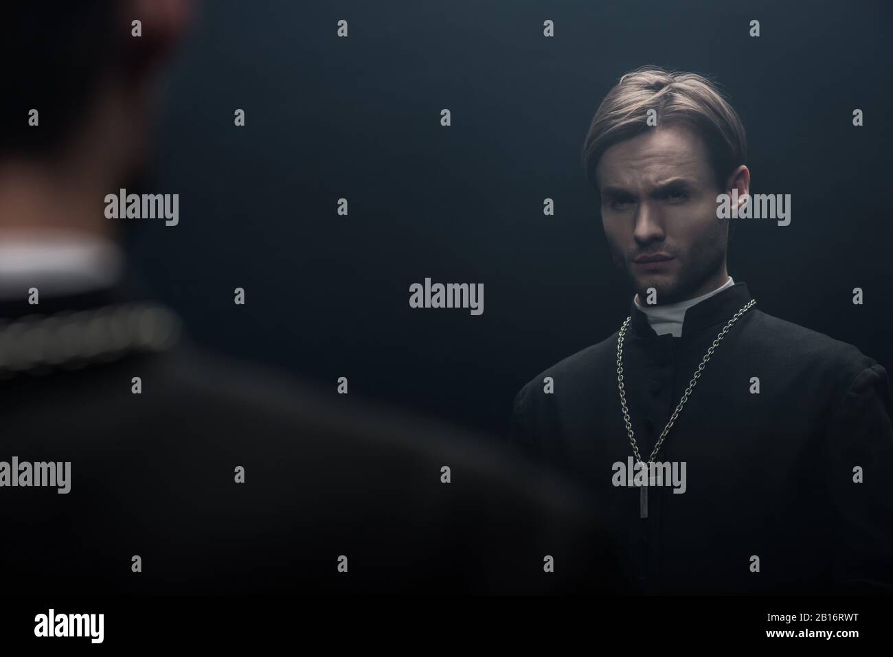 Junger, angespannter, katholischer Priester, der die eigene Reflexion auf Schwarz betrachtet Stockfoto