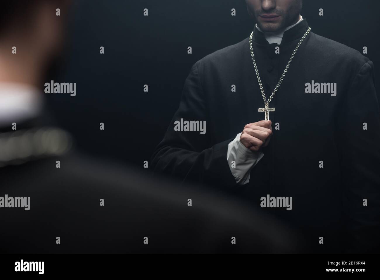 Beschnittenes Bild des katholischen Priesters, der das Kreuz auf seiner Halskette nahe der eigenen Reflexion berührt, isoliert auf Schwarz Stockfoto