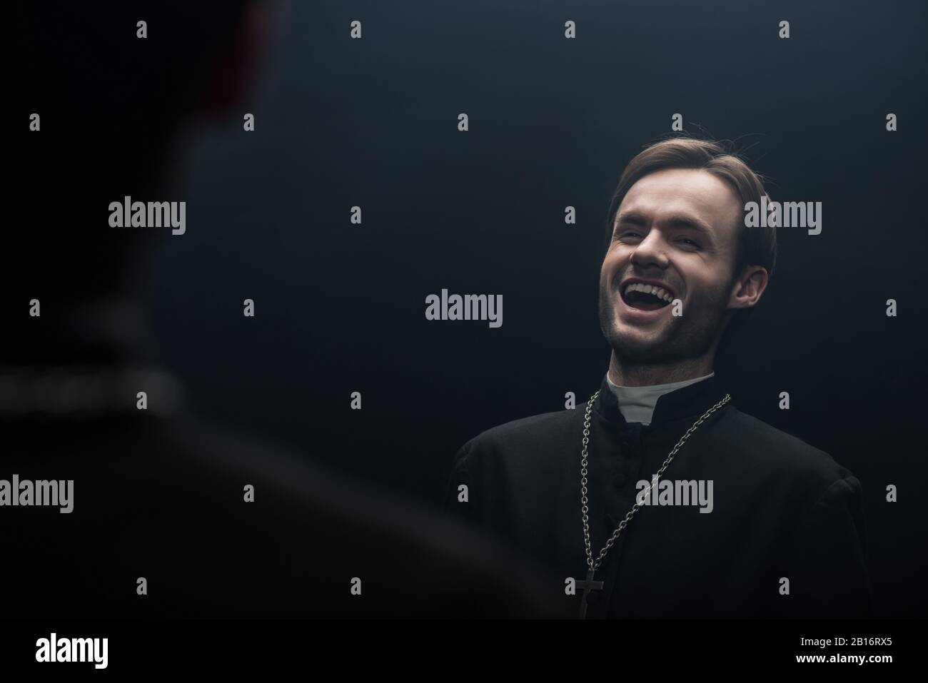 Der junge katholische Priester lacht sarkastisch, während er seine eigene Reflexion betrachtet, die auf Schwarz isoliert ist Stockfoto