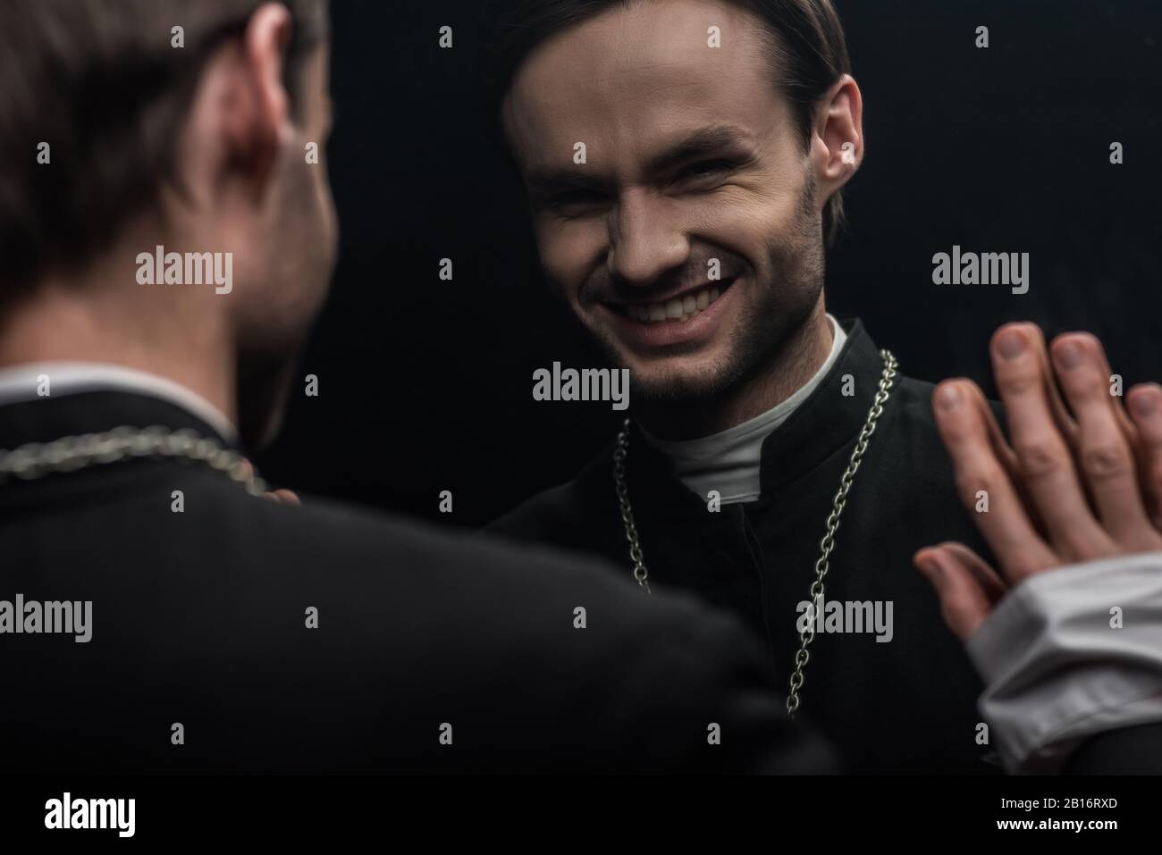 Der junge katholische Priester lacht sarkastisch, während er seine eigene Reflexion betrachtet, die auf Schwarz isoliert ist Stockfoto