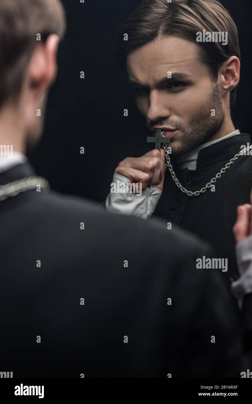 Der junge, angespannte katholische Priester küsst das heilige Kreuz, während er seine eigene Reflexion betrachtet, die auf Schwarz isoliert ist Stockfoto
