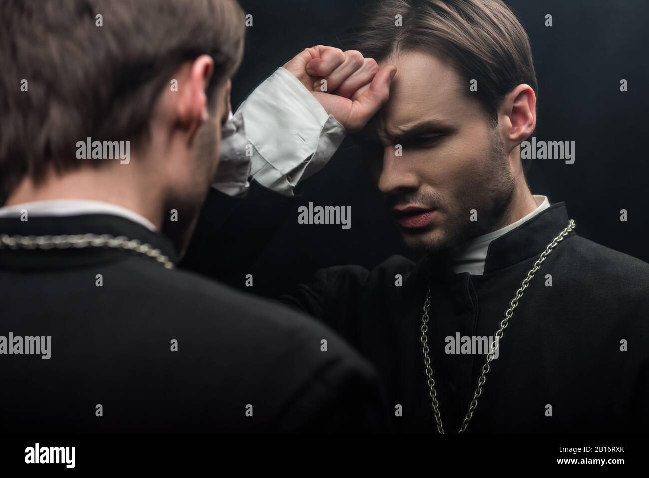 Junger, angespannter, katholischer Priester, der die Faust in der Nähe der Stirn hält, während er in der Nähe der eigenen Reflexion steht, isoliert auf Schwarz Stockfoto