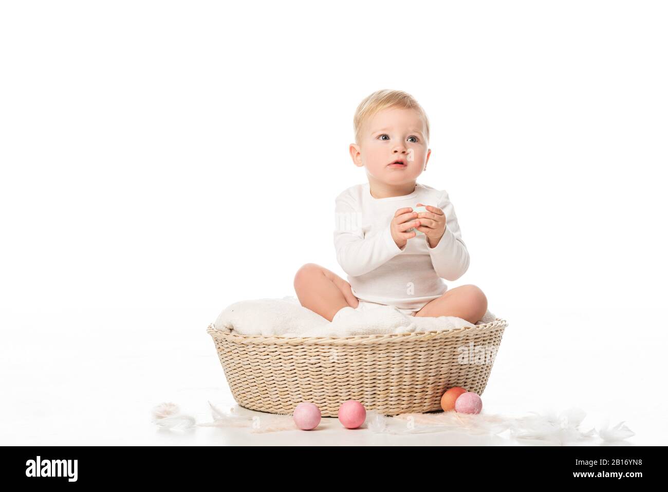 Kind, das Osterei hält, sitzt in einem Korb mit farbenfroher Dekoration auf weißem Hintergrund Stockfoto