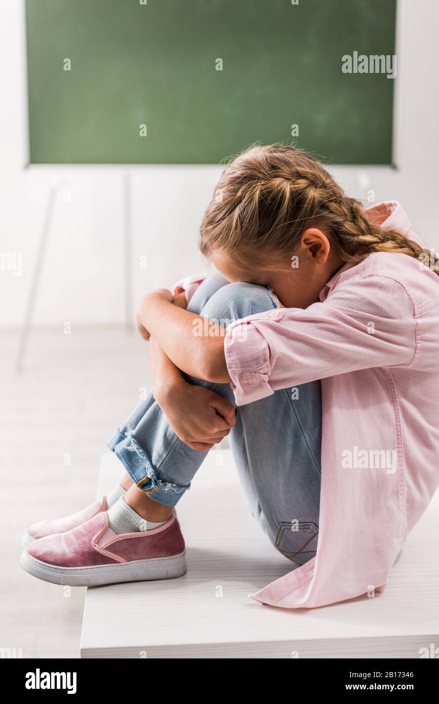 Verärgert und drangsaliert Schulmädchen, die im Klassenzimmer auf dem Schreibtisch sitzen Stockfoto